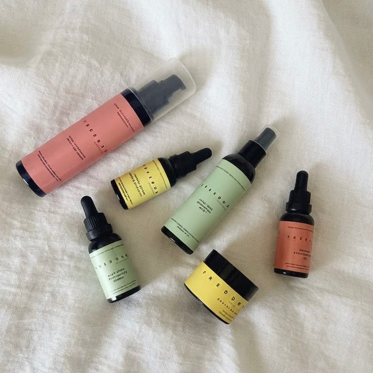 Freedom Self-Love 100% natural waterless skincare collection featuring balms, face oils, and mists in dark glass packaging arranged on a textured neutral textile.